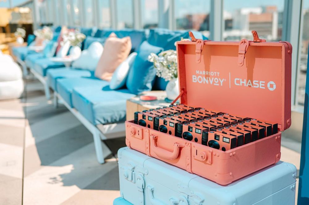 Coral Marriott Bonvoy and Chase suitcase holding branded disposable cameras for guests, beside blue poolside lounges